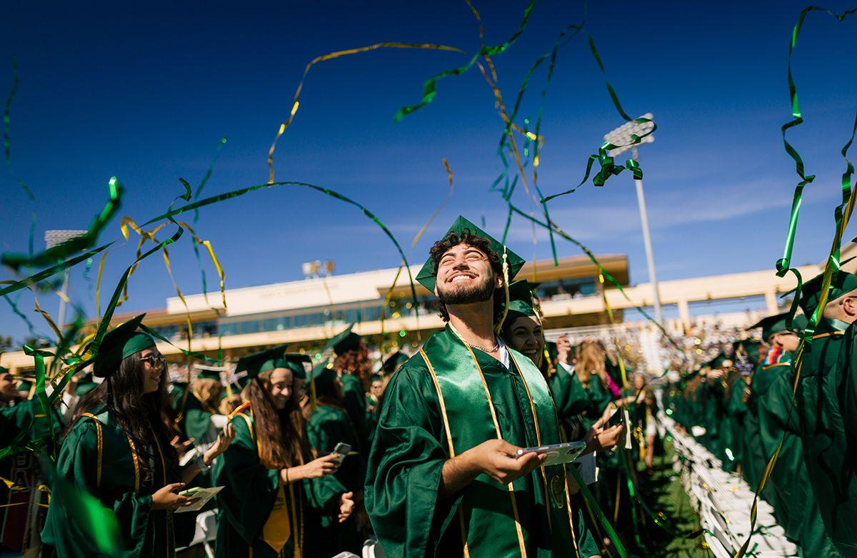 Graduates in green caps and gowns celebrating outdoors with green and gold streamers in the air on a sunny day.