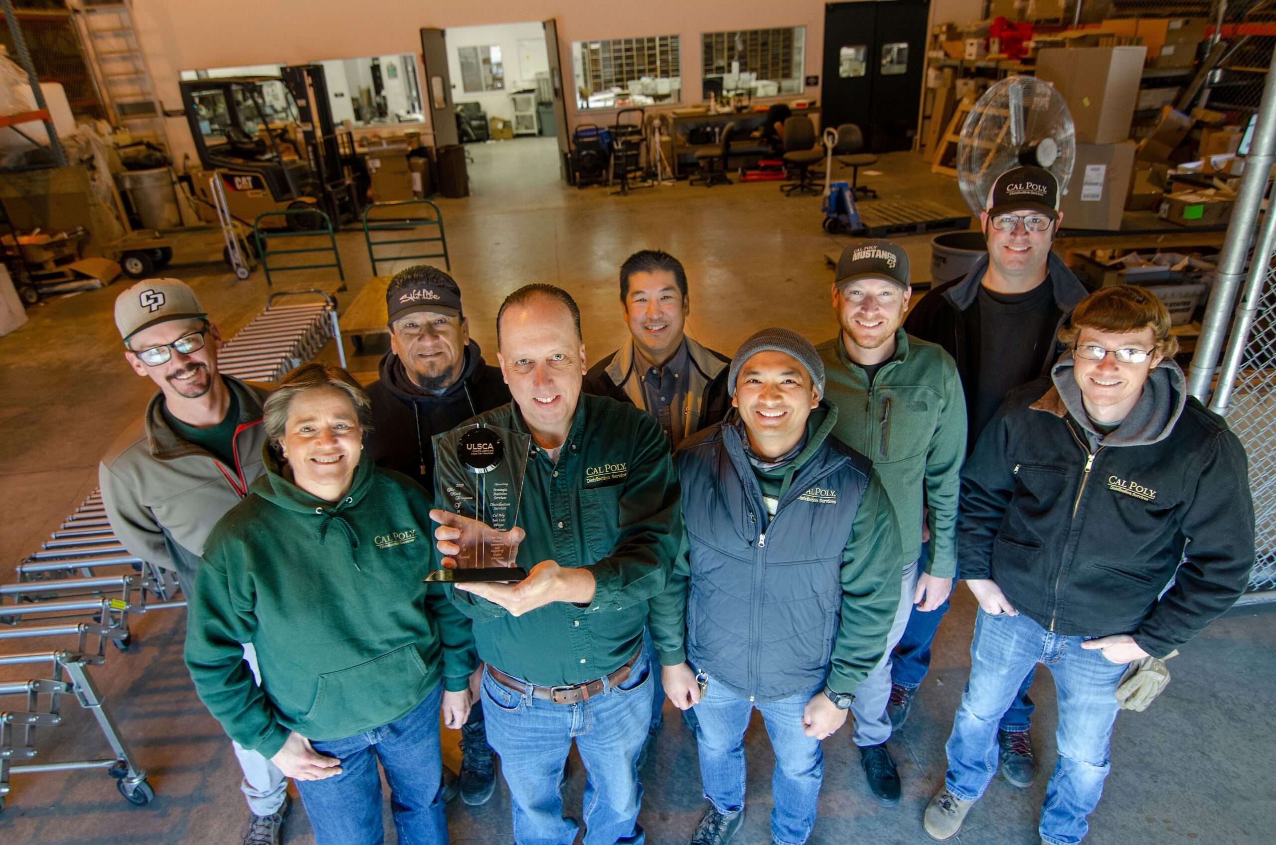Group of nine people smiling in a warehouse, one holding a ULSCA award trophy. Many wear Cal Poly branded clothing.