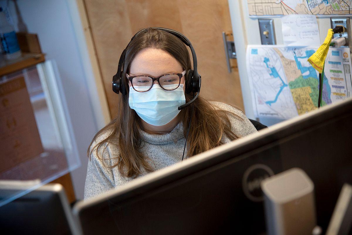 Woman wearing glasses, a face mask, and headset working at a computer in an office with maps on the wall.