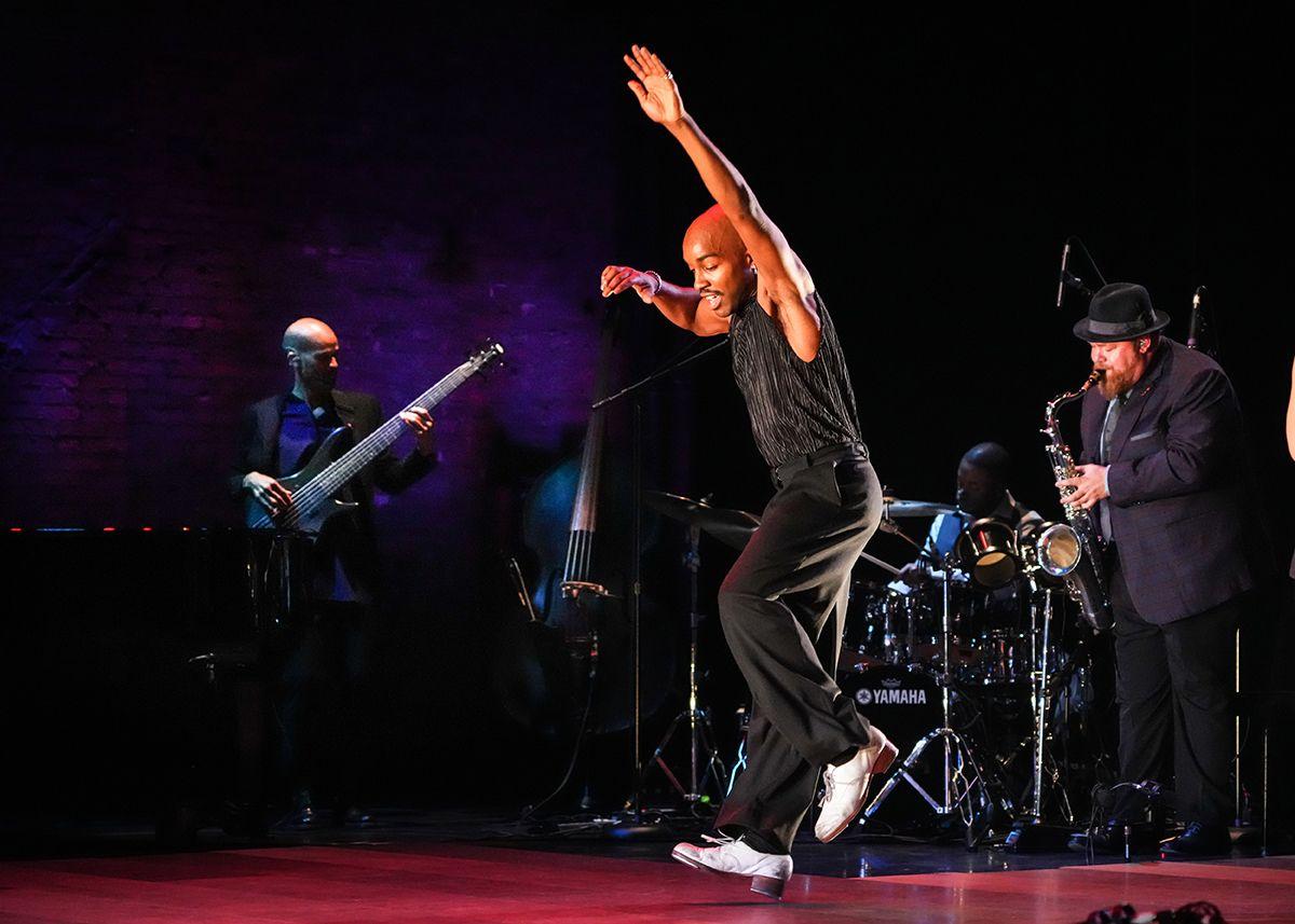 Tap dancer performing energetically on stage with jazz band including bassist, saxophonist, and drummer.
