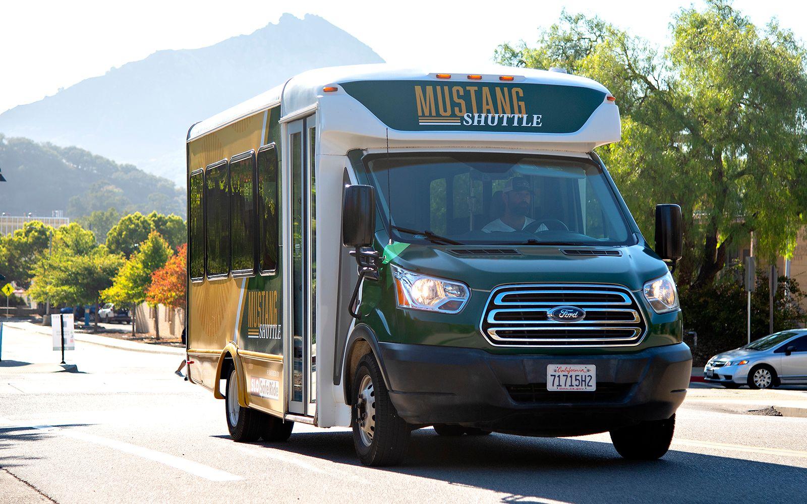 Green and yellow Mustang Shuttle bus driving on a sunny road with mountains and trees in the background.