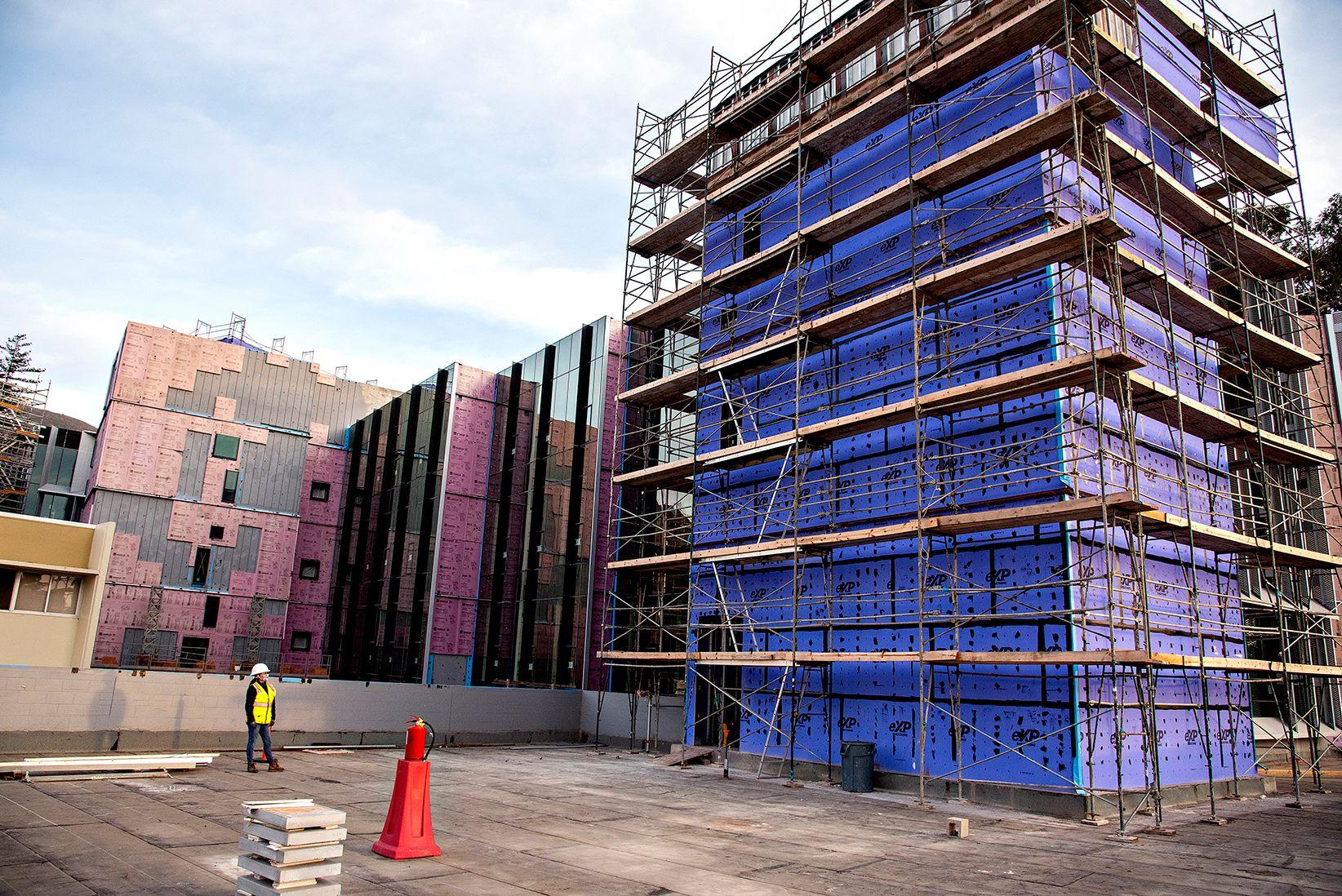 Building under construction with scaffolding, covered in blue and pink insulation panels, worker in safety gear nearby.