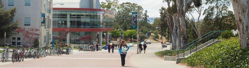 Campus walkway with people walking, bicycles parked, trees, and modern buildings in the background.
