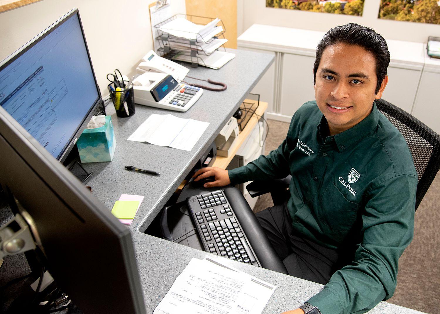 Man in green Cal Poly shirt sitting at an office desk with dual monitors, papers, calculator, and keyboard.