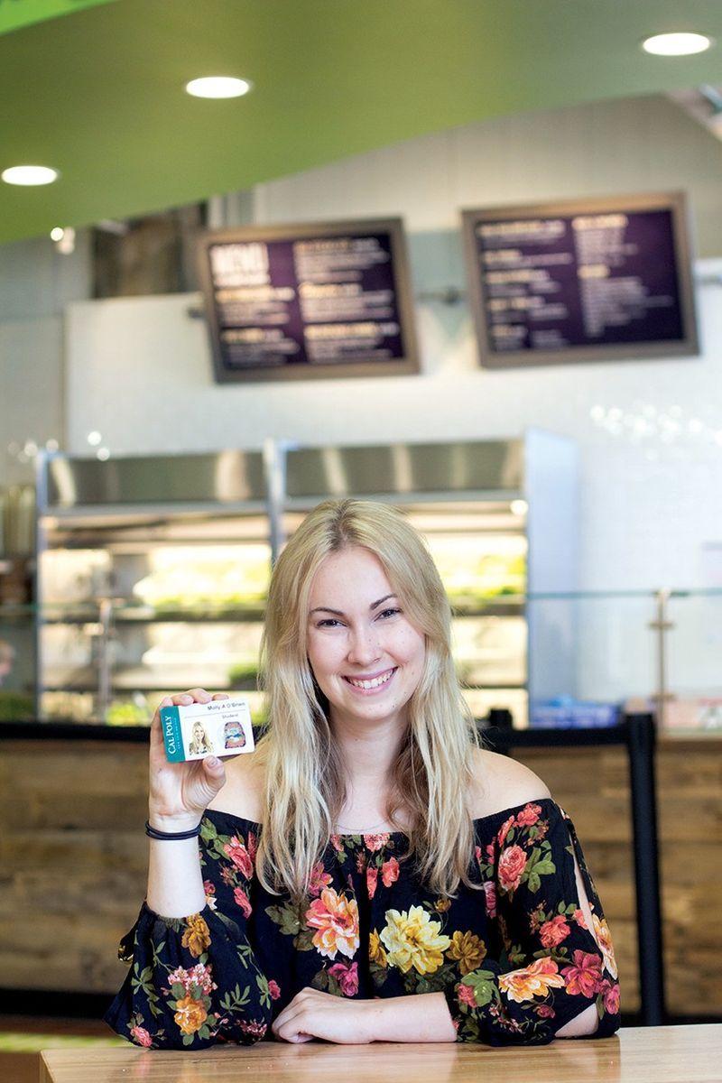 Smiling woman in floral blouse holds Cal Poly student ID card while sitting at a table in a cafe.