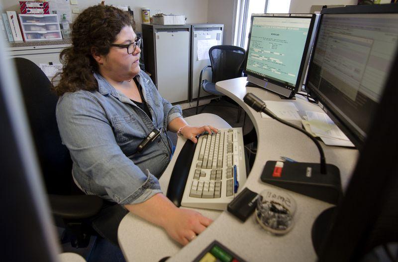 Woman in glasses and denim shirt typing on a keyboard at a desk with dual monitors and a microphone.