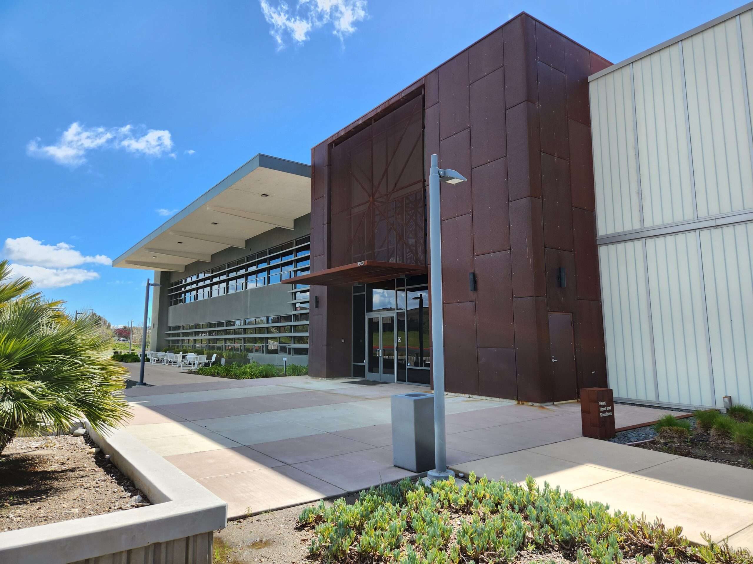 Modern building with rust-colored and gray walls, large windows, outdoor seating, and nearby greenery under a blue sky.