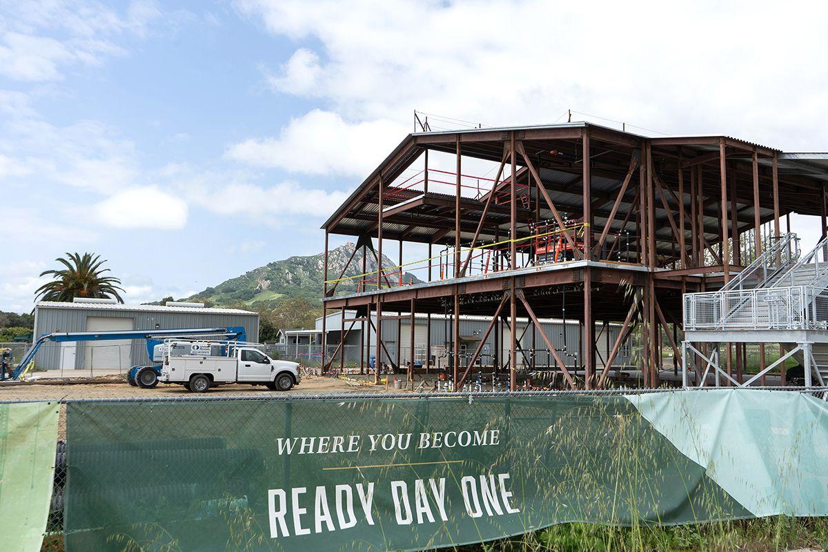 Partially built steel frame structure at a construction site with a green fence sign reading "WHERE YOU BECOME READY DAY ONE.
