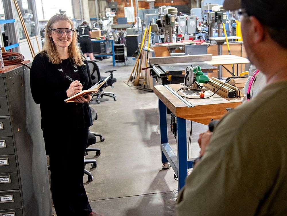 Woman wearing safety goggles writing in a notebook inside a workshop with tools and machinery.