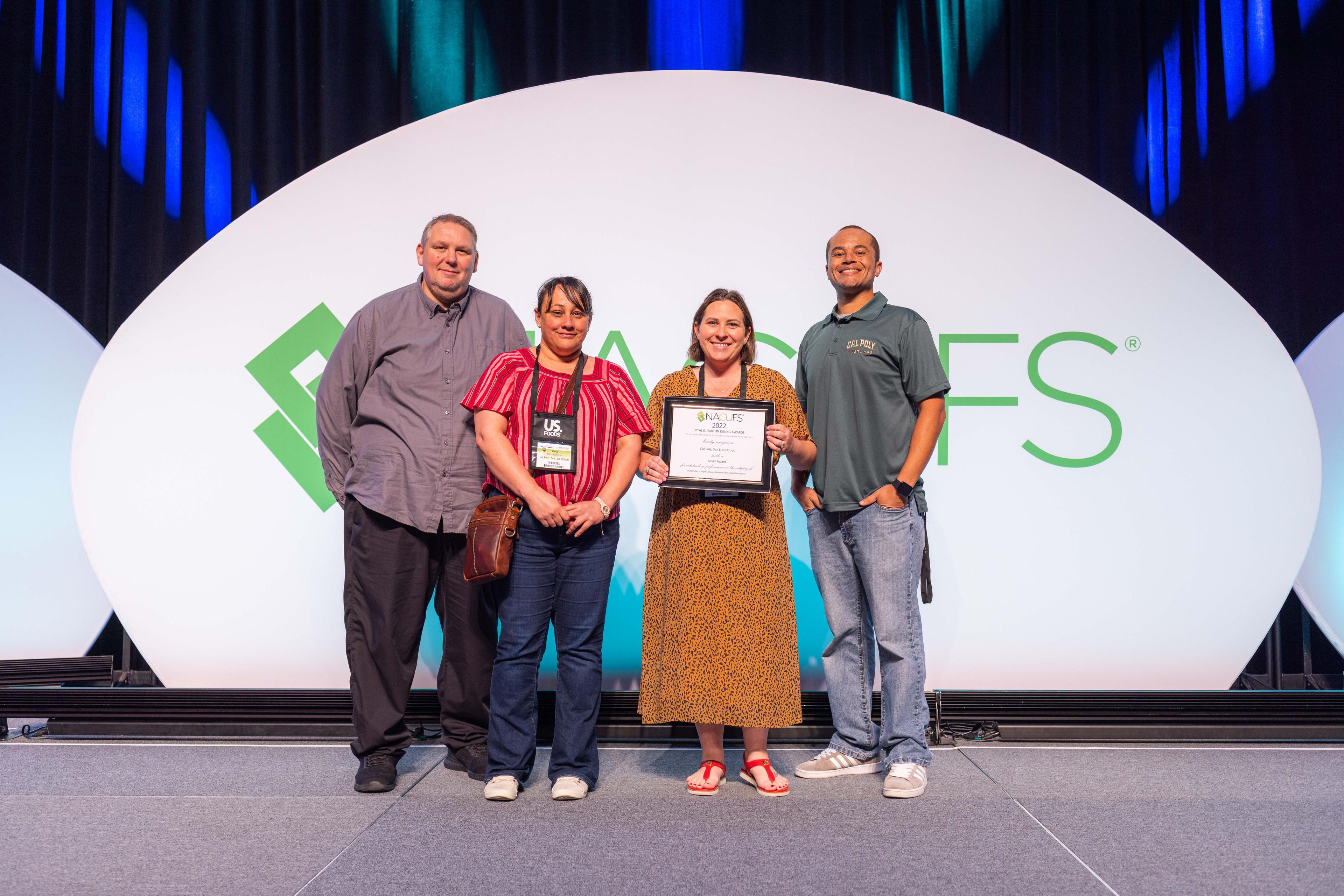Four people standing on a stage in front of a NACUFS logo backdrop, one holding a certificate.