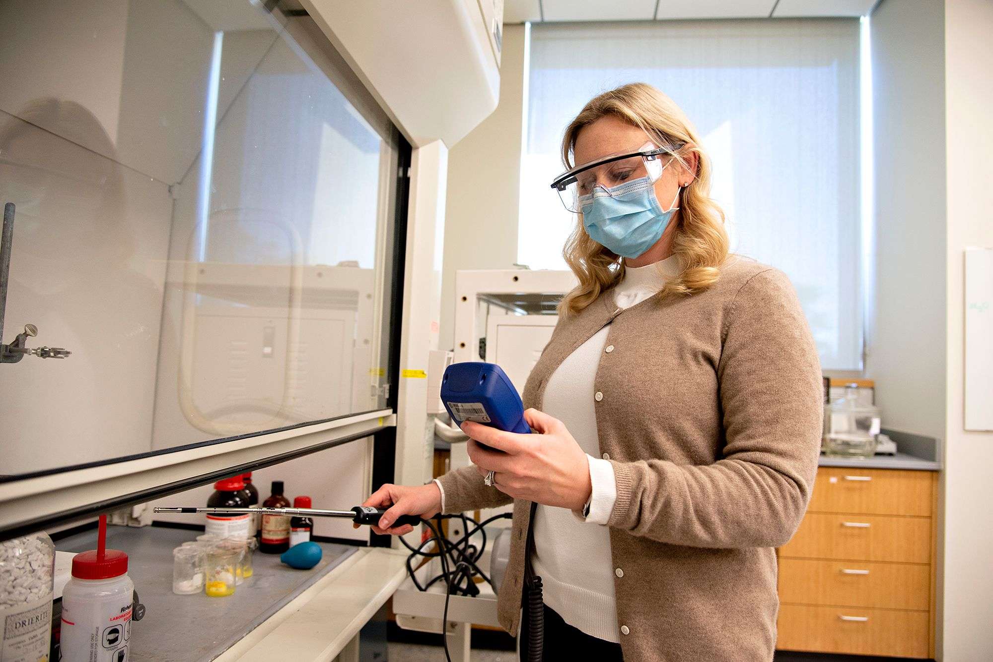 Woman wearing safety glasses and mask operating a device inside a laboratory fume hood.