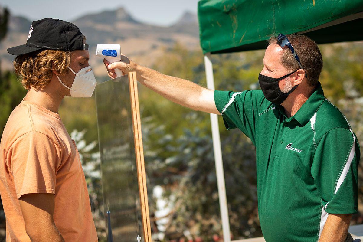 Man in green shirt and black mask checks temperature of masked man in peach shirt and backward cap outdoors.