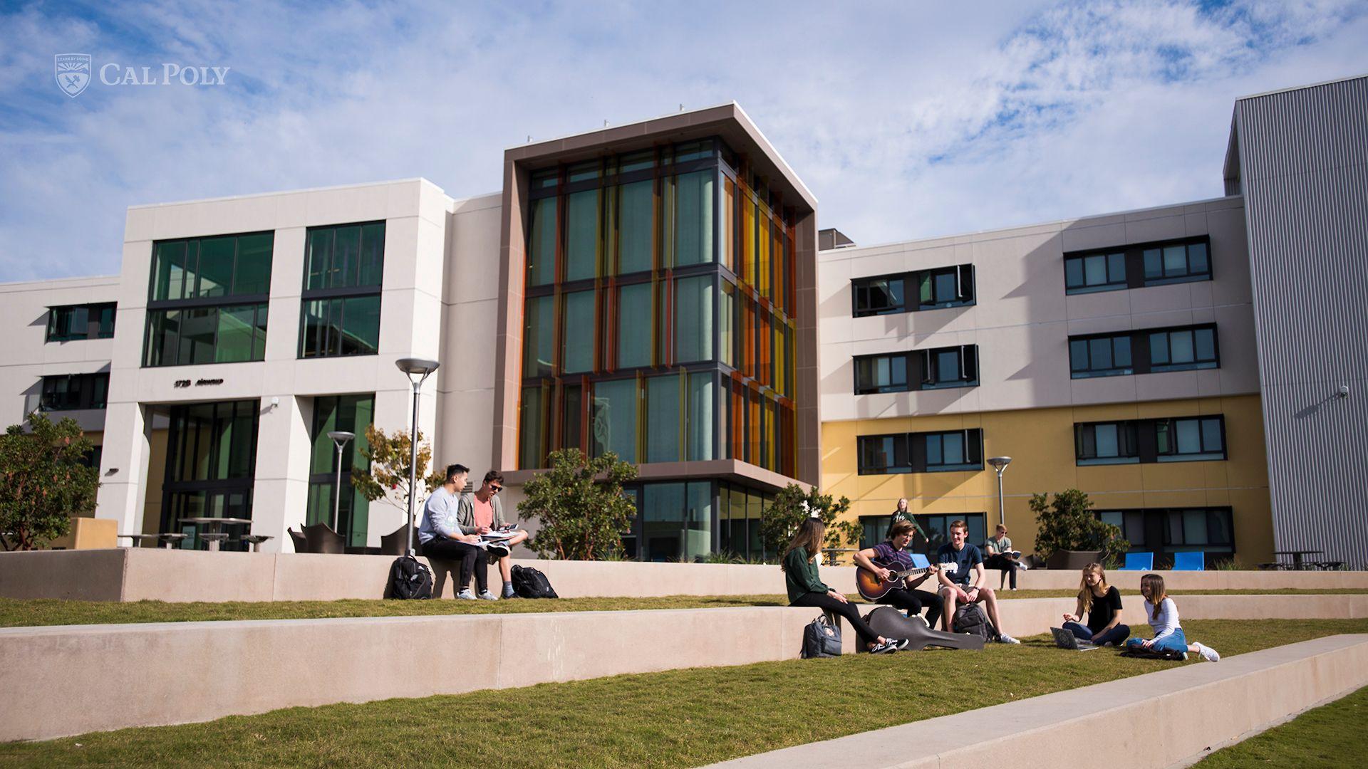 Students sitting and studying on tiered grass near a modern Cal Poly campus building under a blue sky.