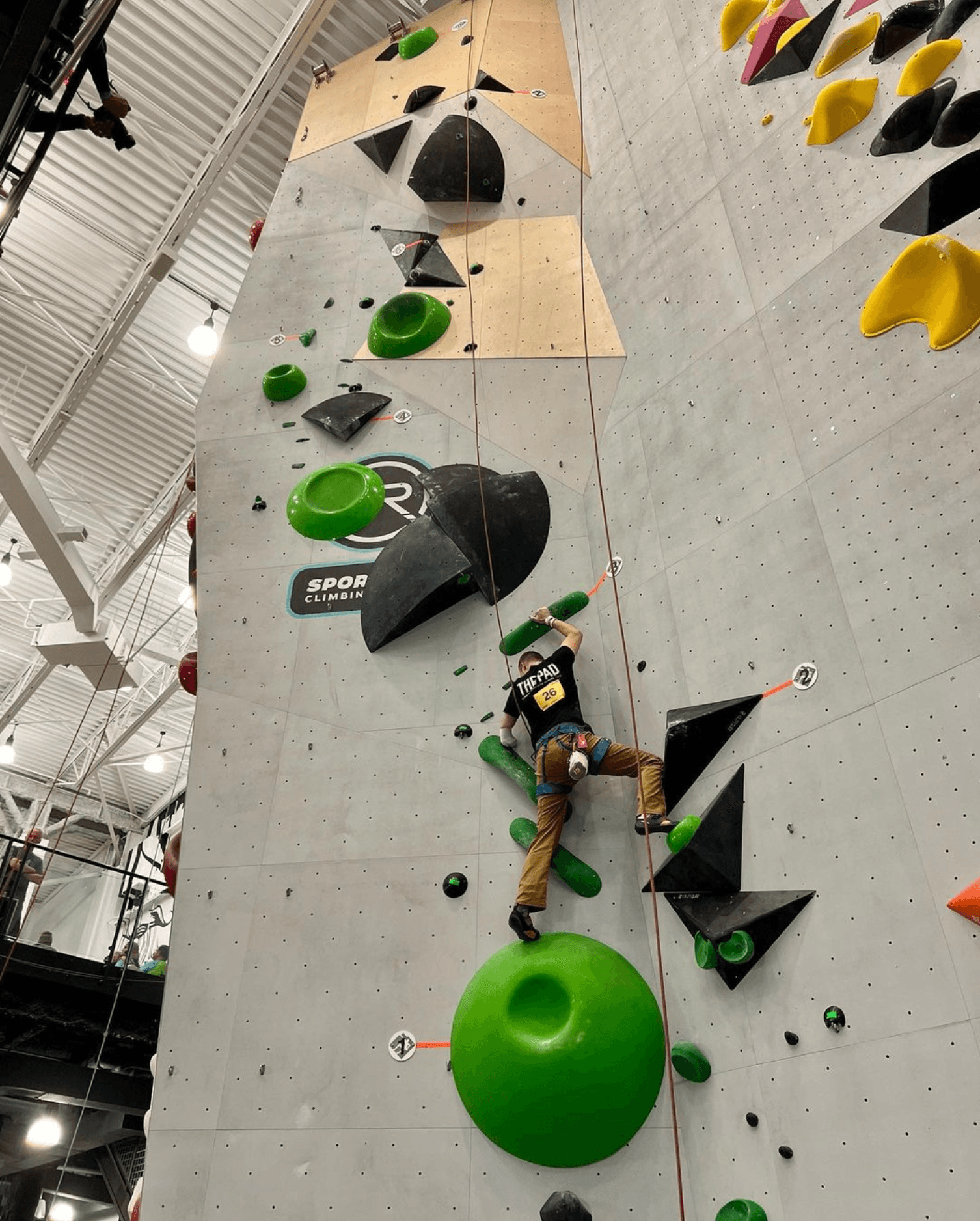 Person climbing an indoor rock wall with large green and black holds, wearing a harness and a "The Pad" shirt.