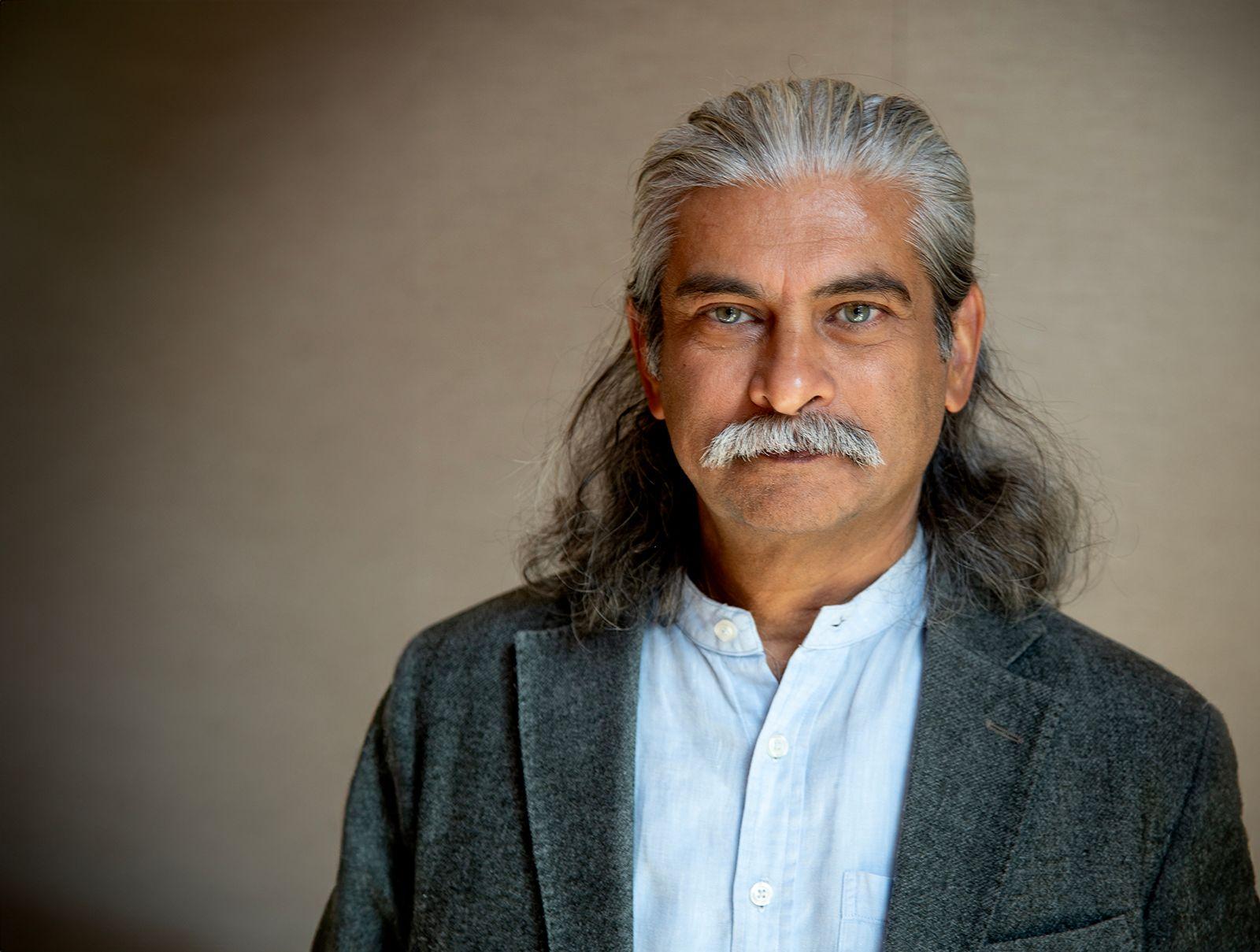 Man with long salt-and-pepper hair, thick mustache, blue eyes, wearing a gray blazer and white buttoned shirt.