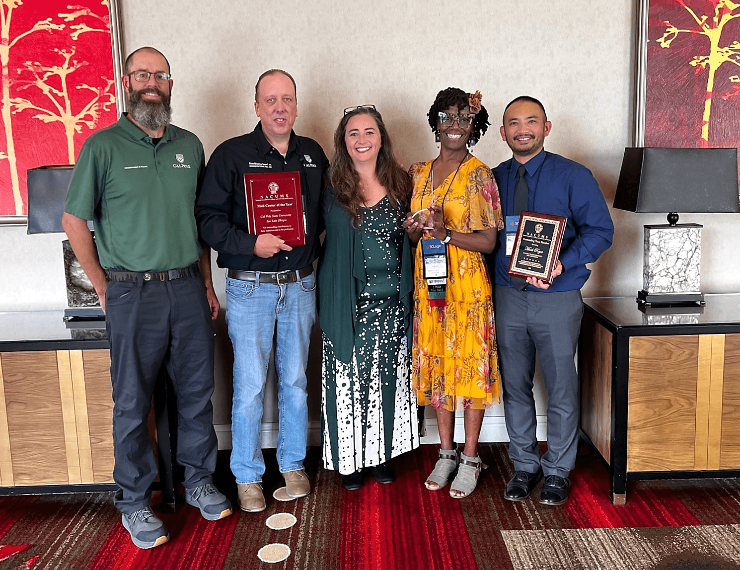 Five people smiling and holding awards, standing between two red artworks and lamps on wooden cabinets.