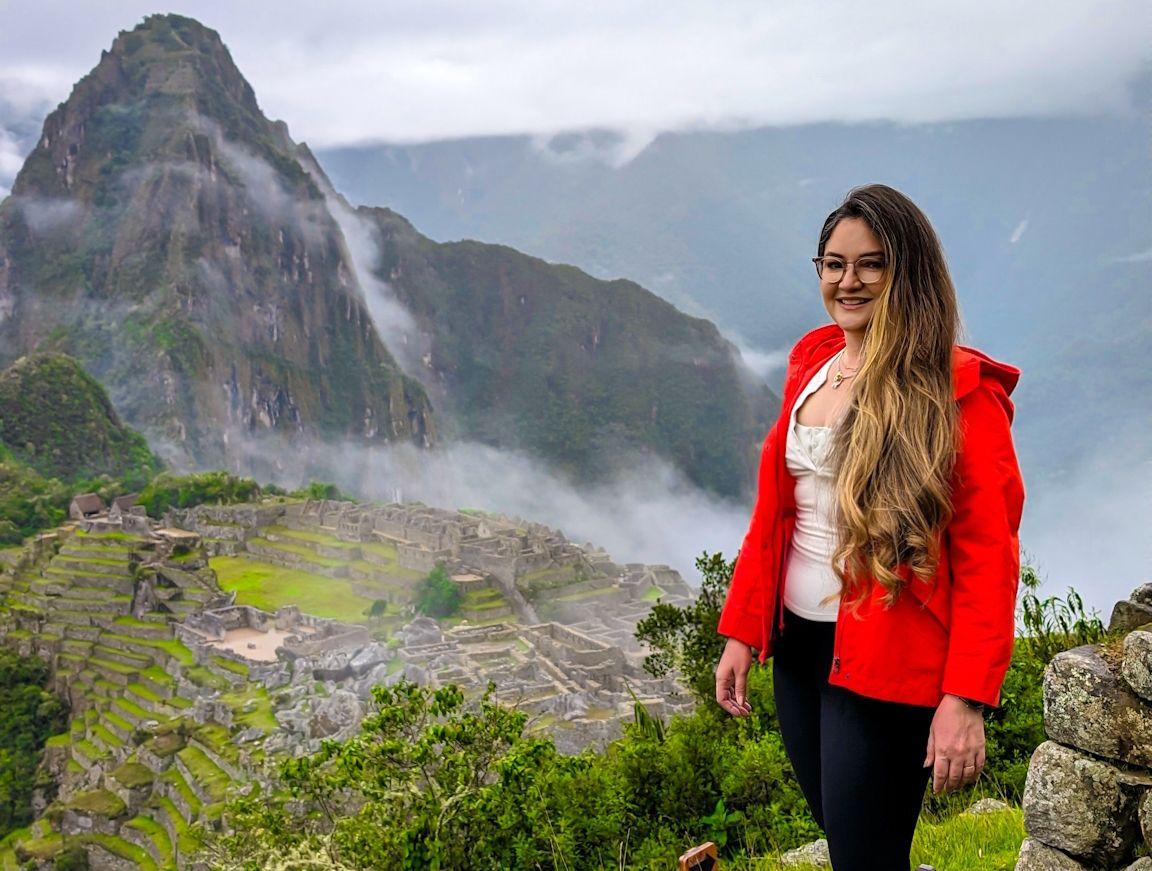 Woman in a red jacket standing near Machu Picchu ruins with mountains and mist in the background.