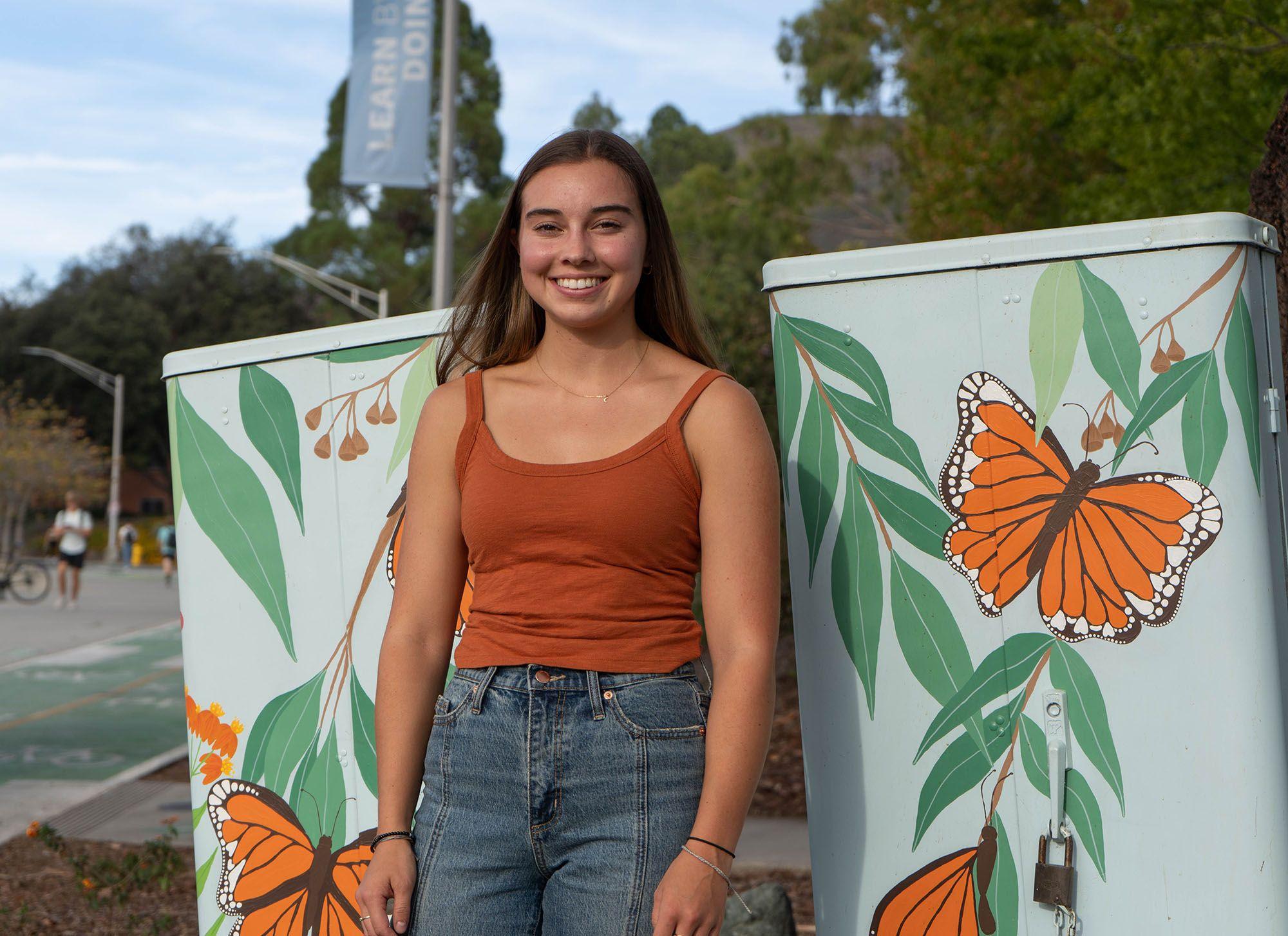Smiling young woman in an orange tank top and jeans standing between two utility boxes painted with orange butterflies and green leaves.