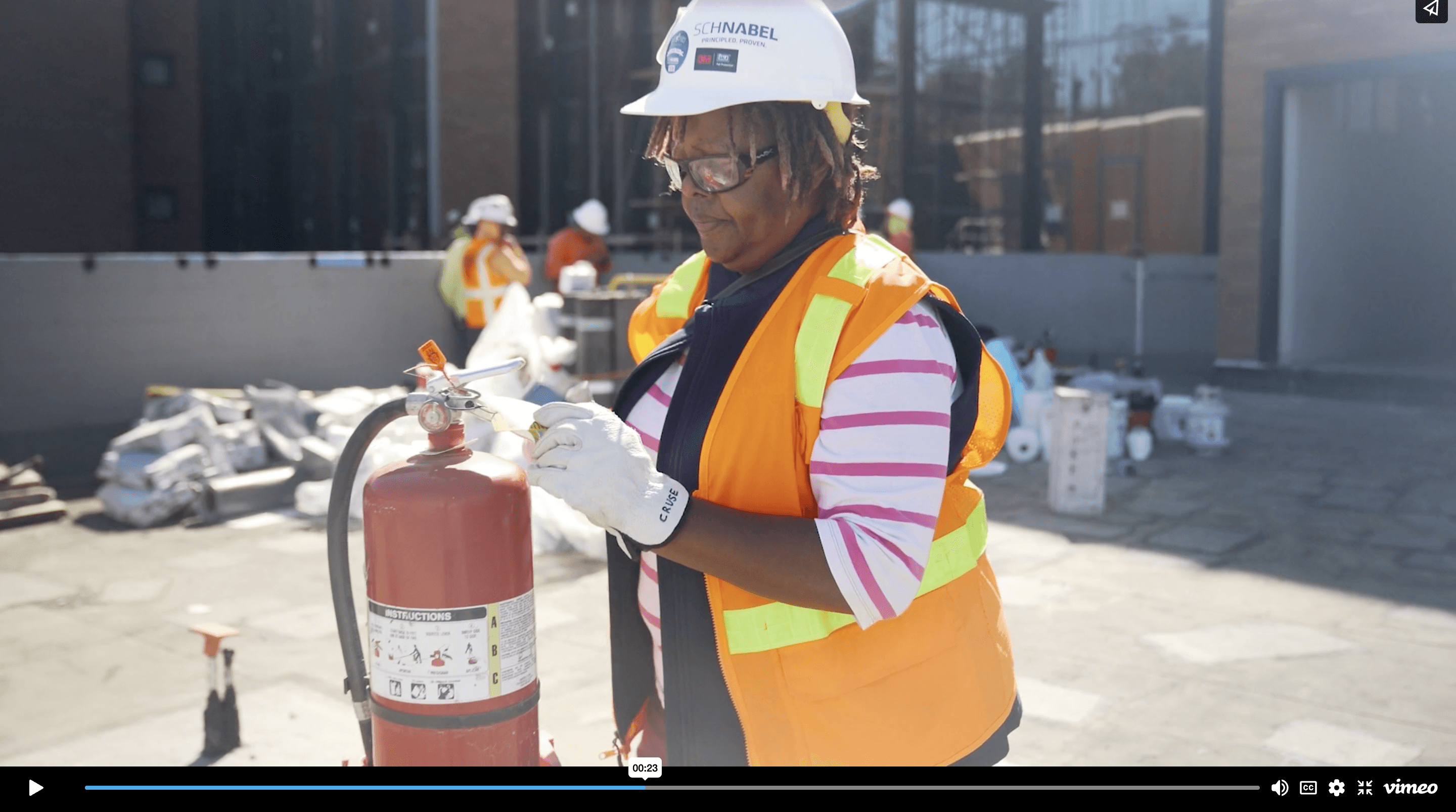 Worker in safety gear inspecting fire extinguisher on construction site with others in background.