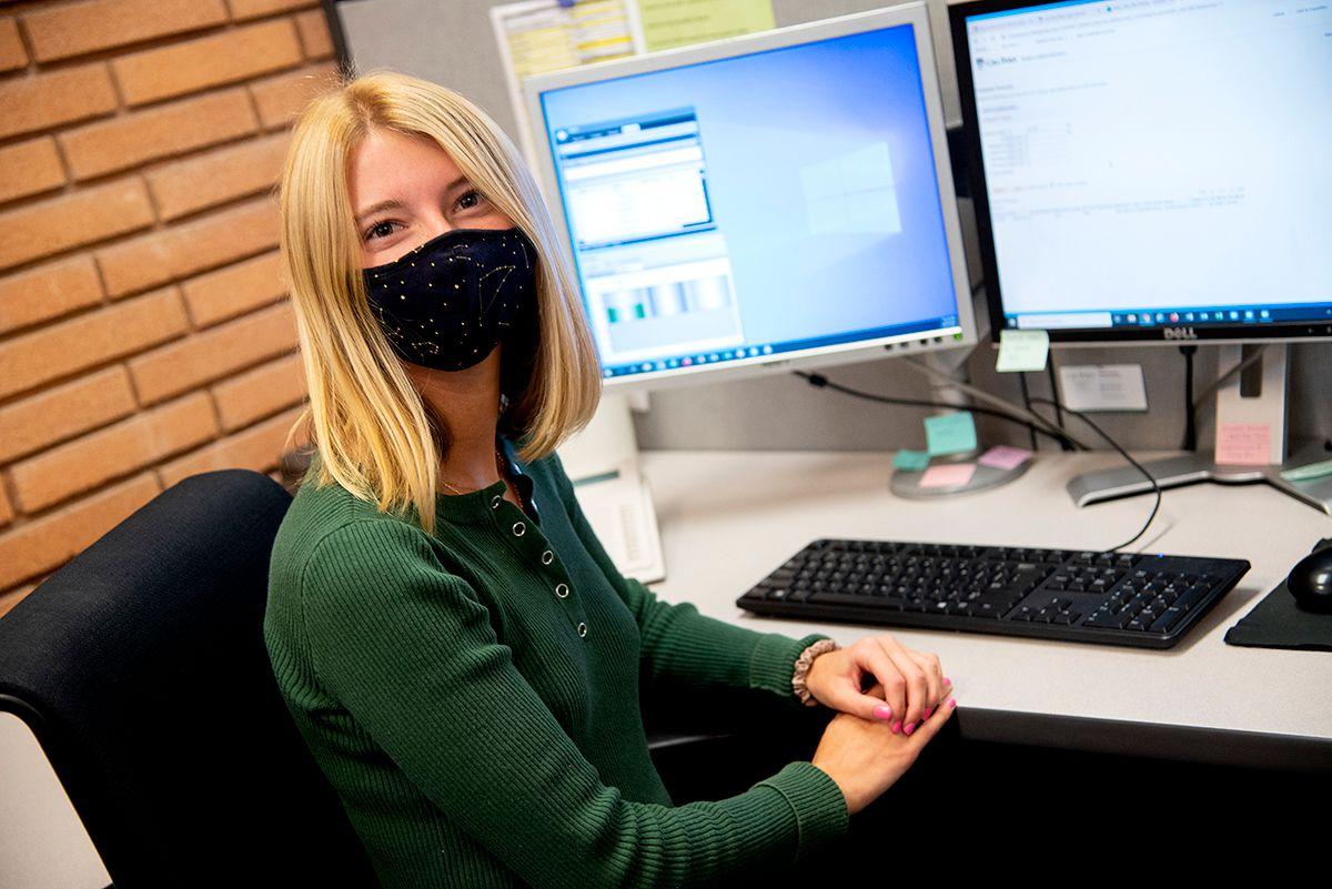 Woman with blonde hair and dark constellation mask sitting at a desk with dual computer monitors and keyboard.