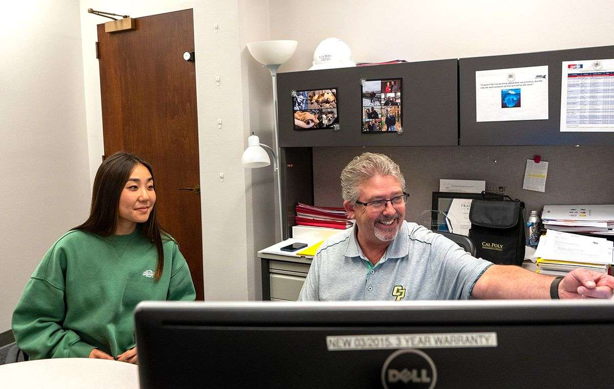 Man with glasses pointing at computer screen, woman in green sweatshirt watching, office setting with photos on wall.