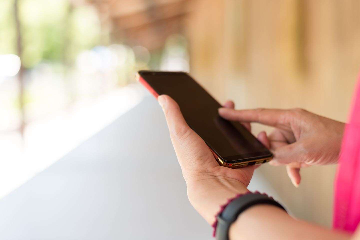 Hand holding a smartphone with the other hand touching the screen, blurred background, and person wearing a fitness tracker.
