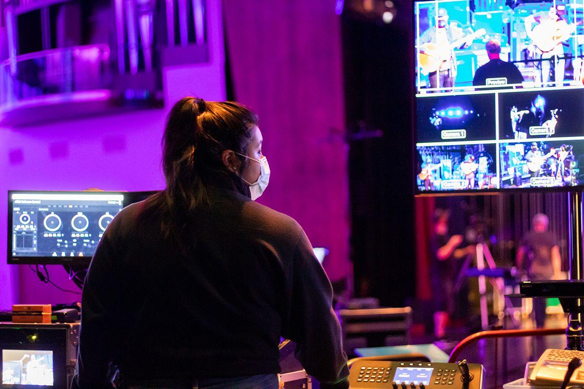 Person wearing a mask operating video equipment with multiple screens showing a live performance in a purple-lit control room.