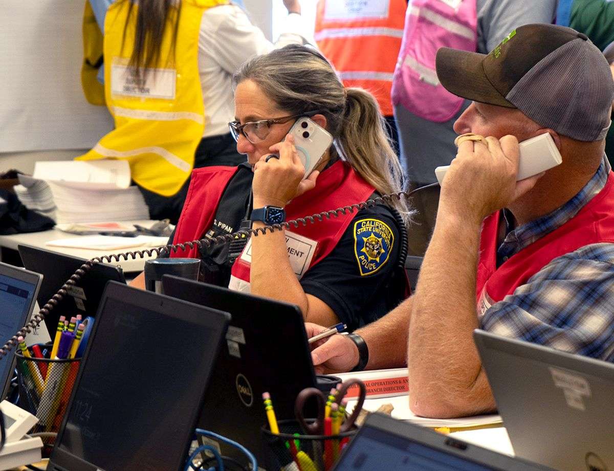 Two emergency responders wearing red vests use phones while working at laptops in a busy office.