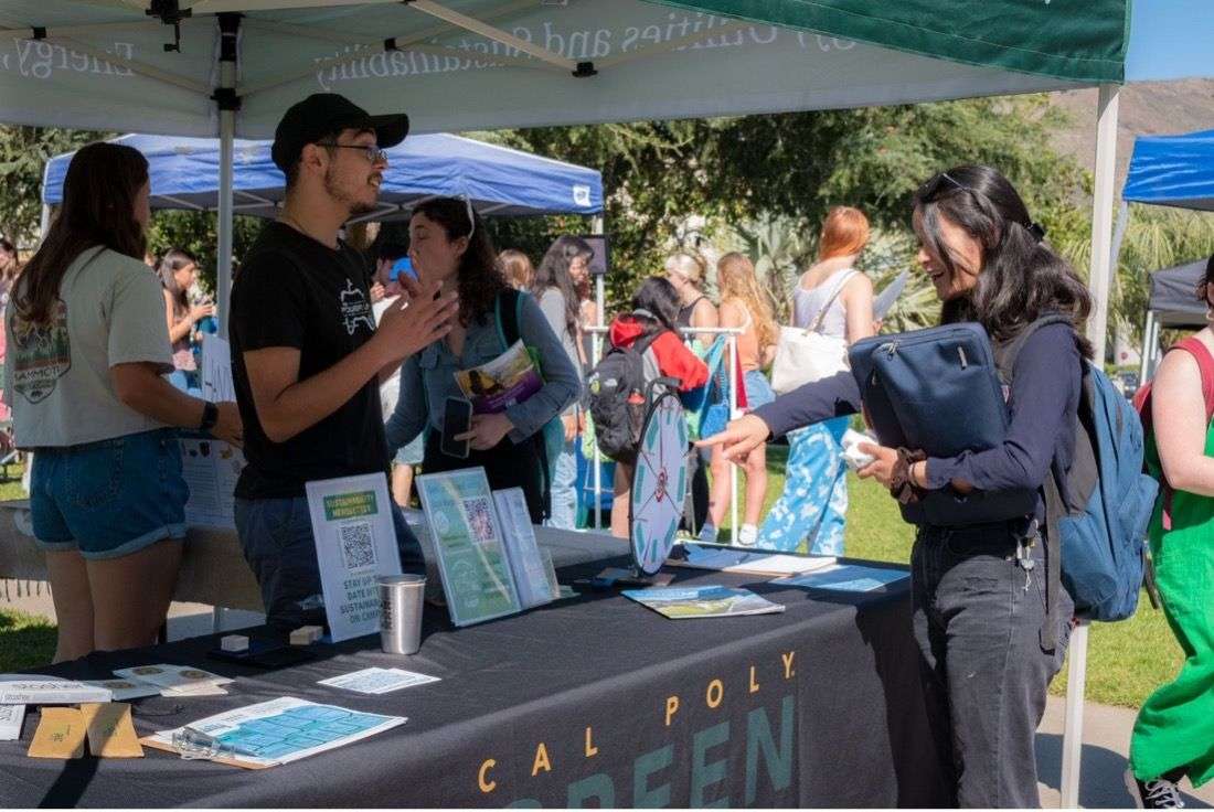 People interacting at a Cal Poly Green booth with a prize wheel at an outdoor event on a sunny day.
