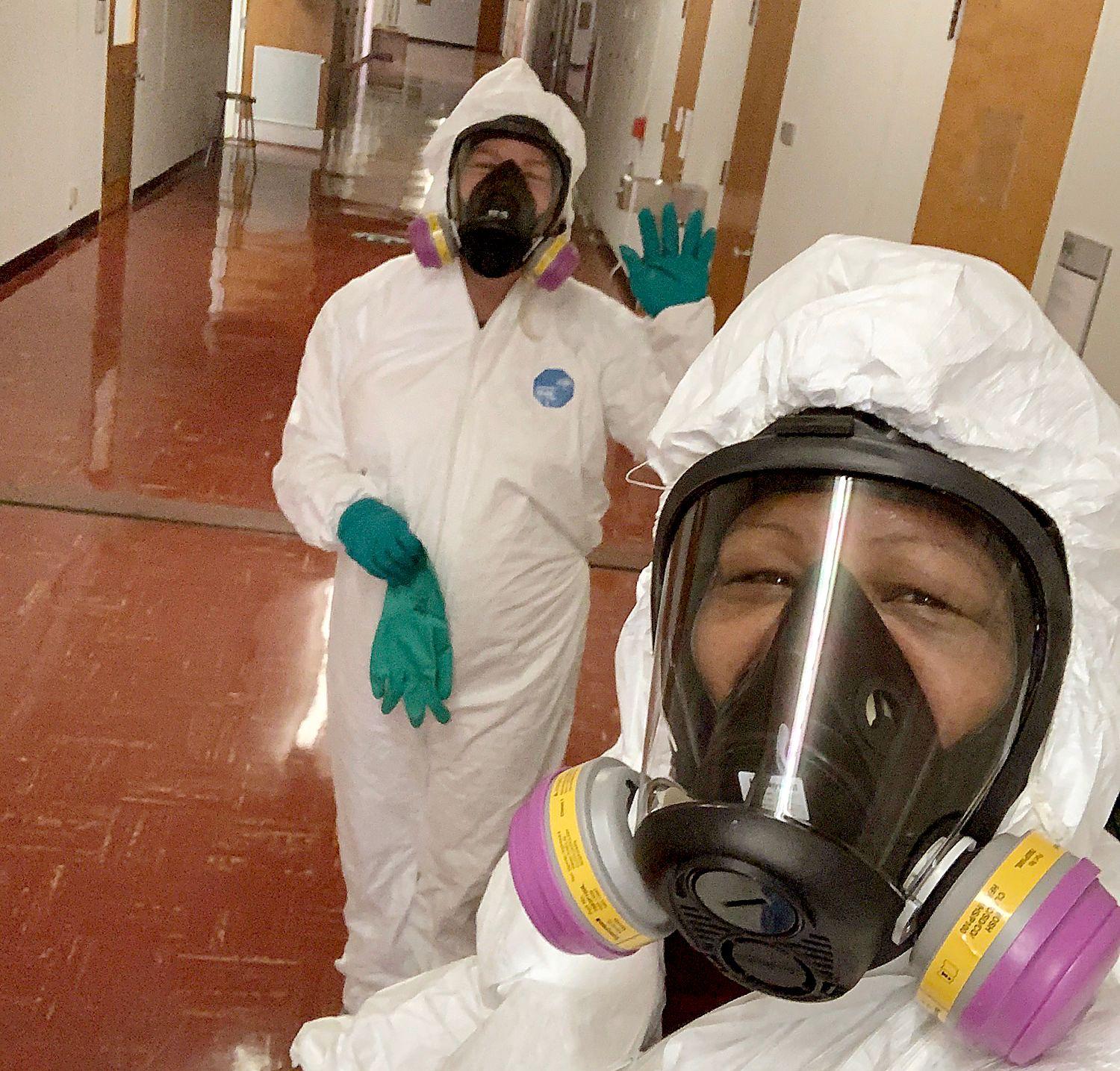 Two people in white hazmat suits, protective masks, and green gloves in a hallway with brown floor tiles.
