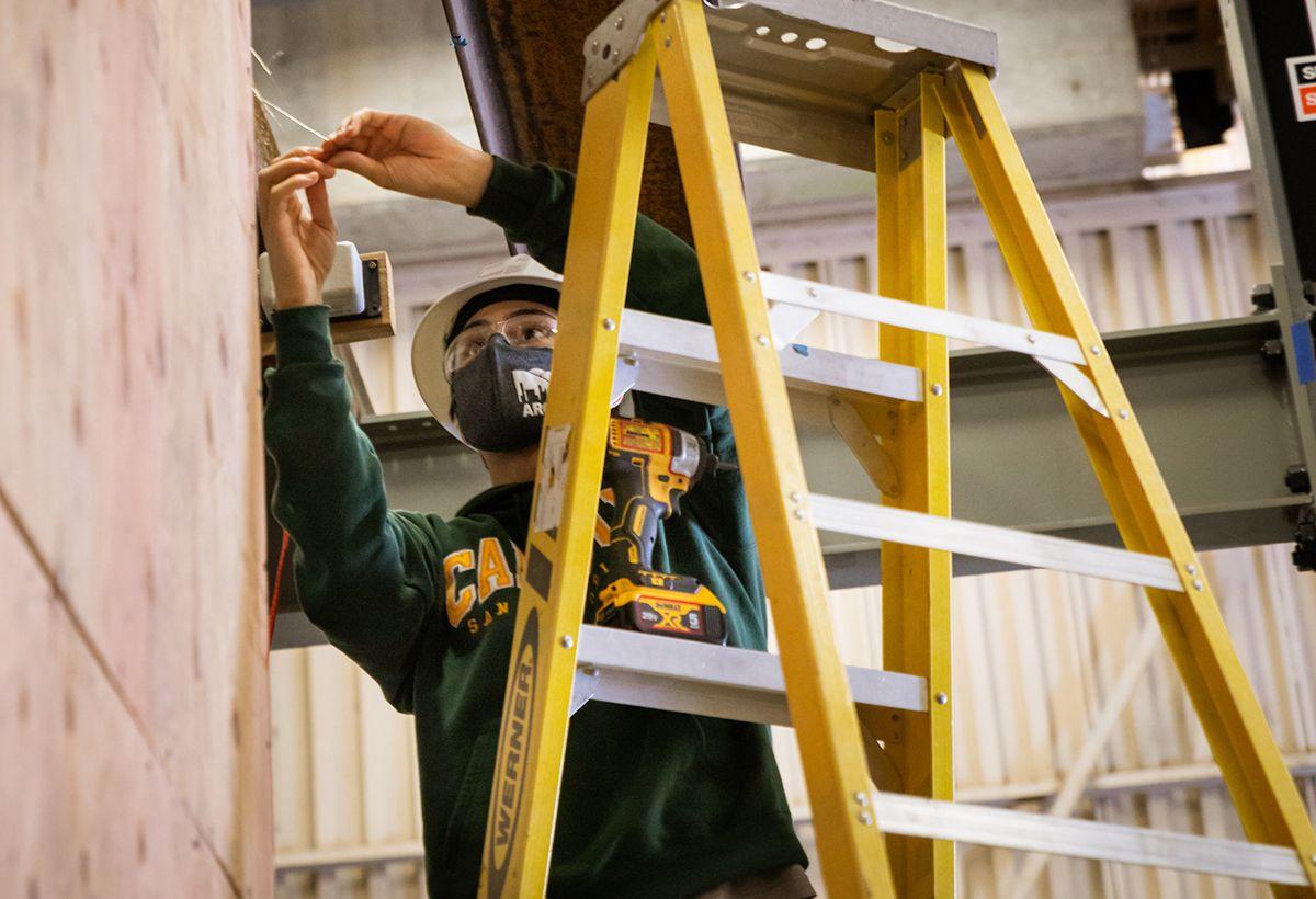 Person wearing safety glasses and mask working on wiring on a yellow ladder in an industrial setting.