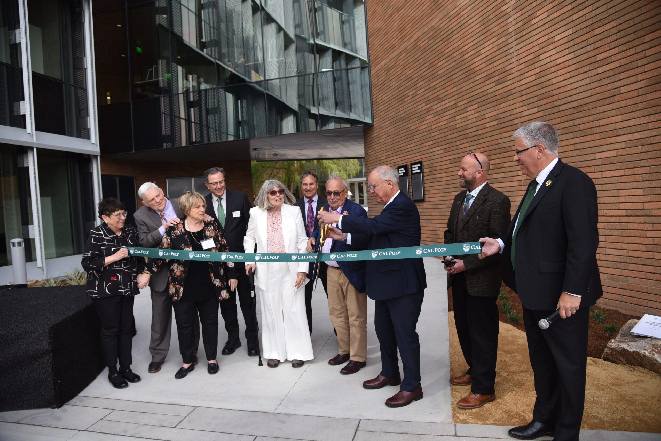 Group of people at a ribbon-cutting ceremony holding a green "Cal Poly" ribbon outside a modern building.