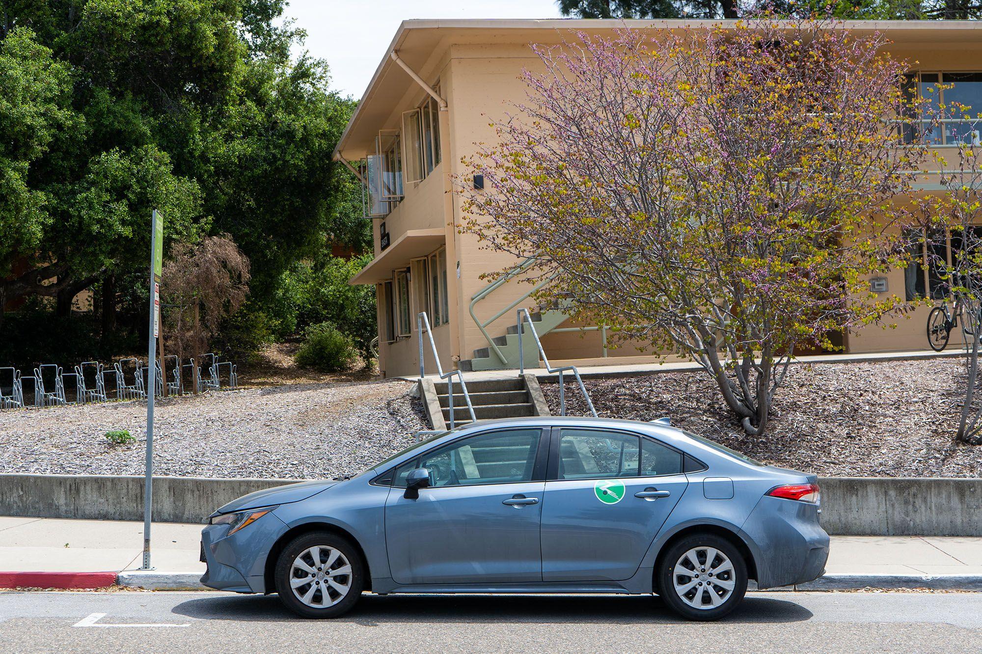 Blue sedan parked by a curb with a green and white car-sharing logo on the rear door, near a beige building and trees.