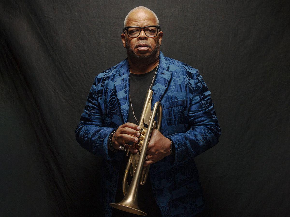 Man wearing glasses and a blue patterned jacket holding a brass trumpet against a dark backdrop.