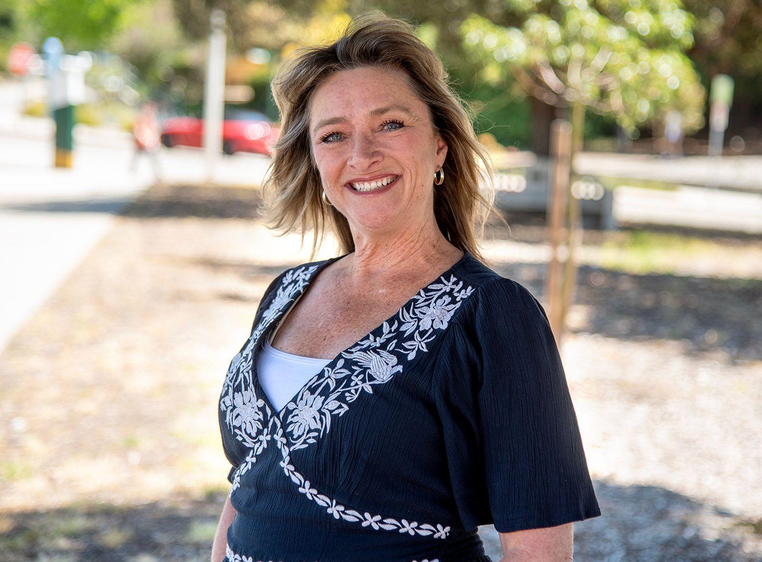 Smiling woman with shoulder-length blonde hair wearing a black top with white floral embroidery outdoors.