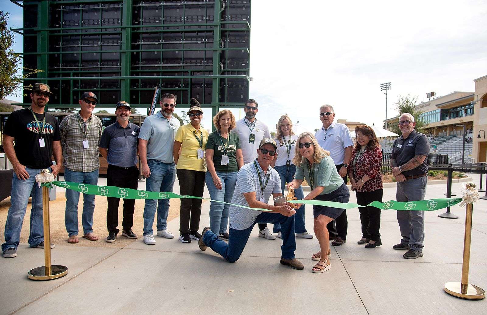 Group of people at an outdoor ribbon-cutting ceremony with a green ribbon featuring white logos.