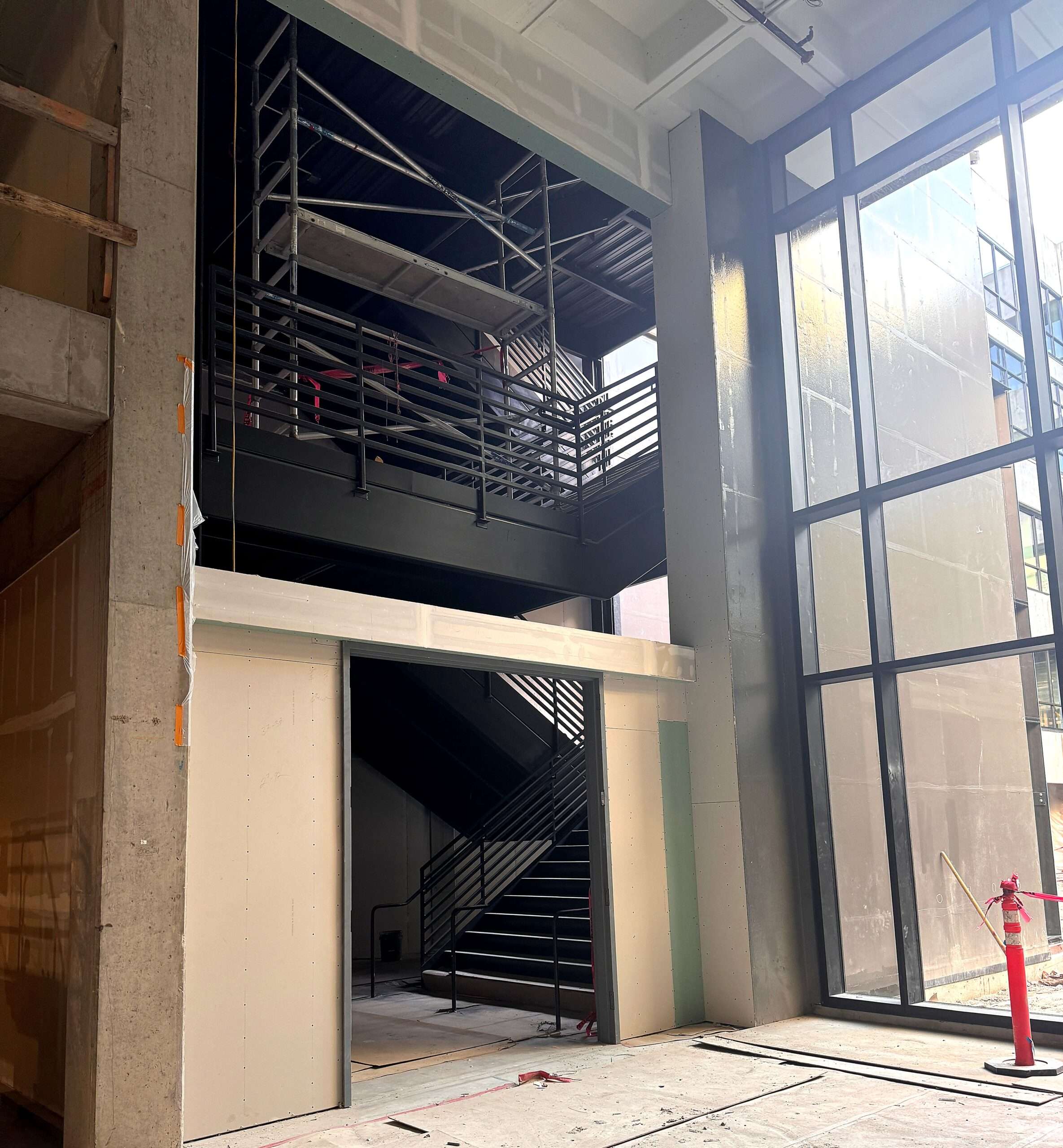 Under-construction building interior with metal staircase, large windows, and scaffolding above the stairs.