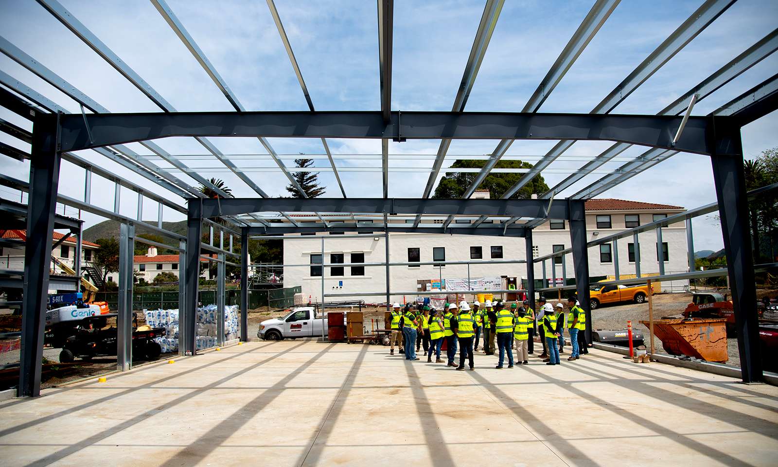 Group of construction workers in yellow vests and hard hats standing inside a steel frame structure under construction.