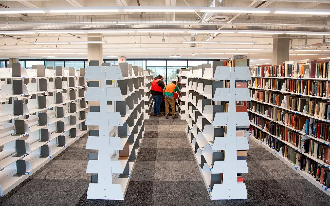 Two people organizing mostly empty white bookshelves in a brightly lit library aisle with one fully stocked shelf.