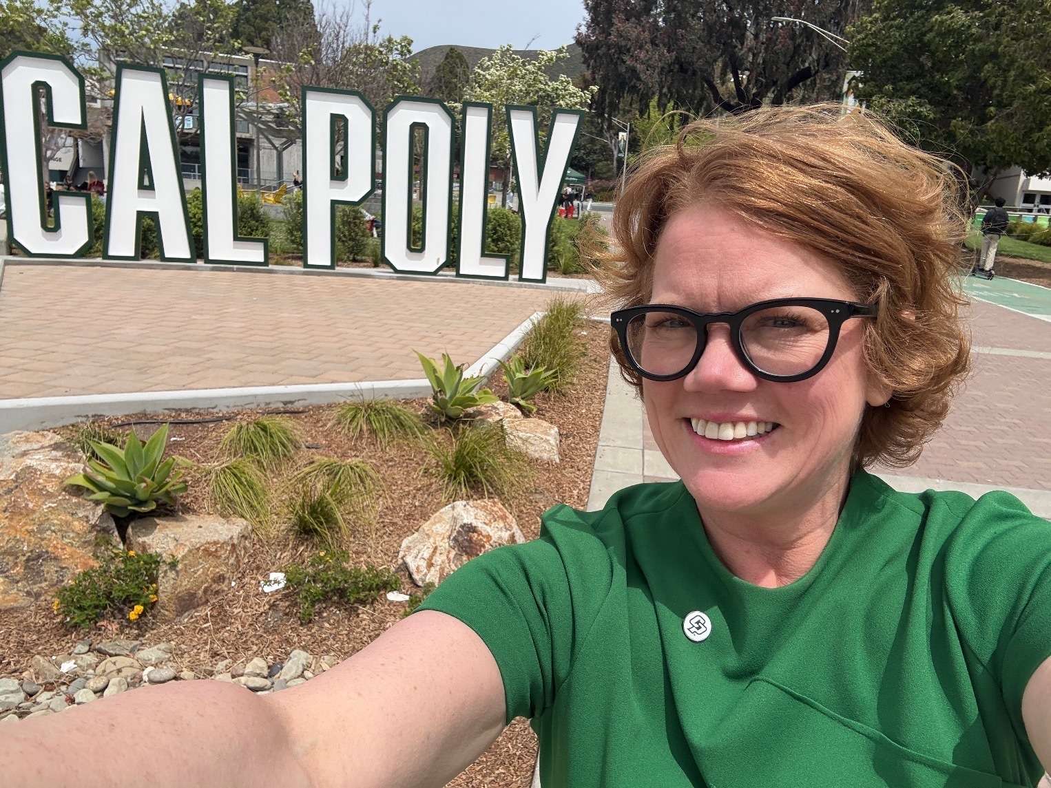 Woman with red hair and glasses smiling for a selfie in front of large CAL POLY letters outdoors.