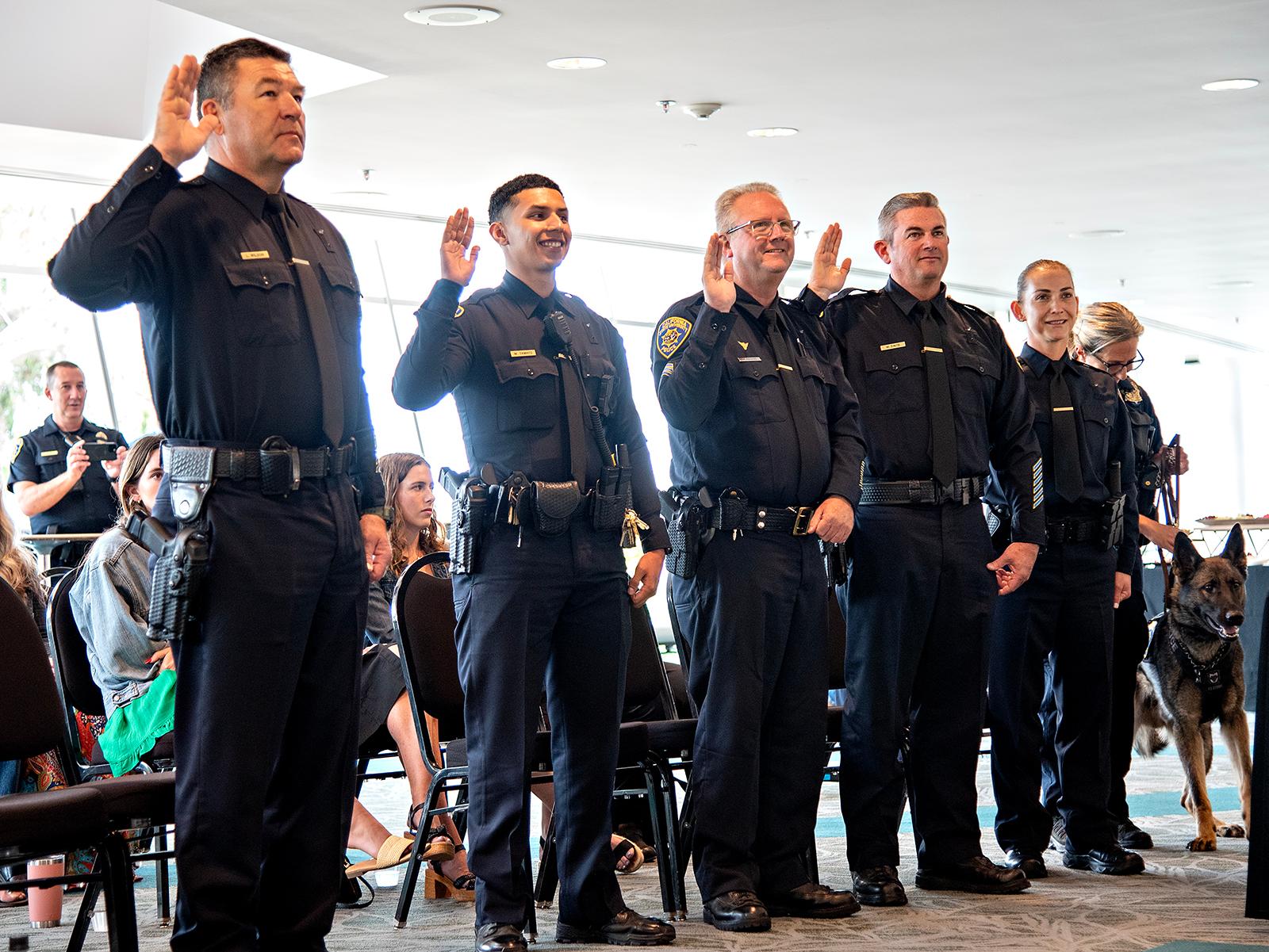 Six uniformed police officers standing with raised right hands, one officer holding a police dog on a leash.