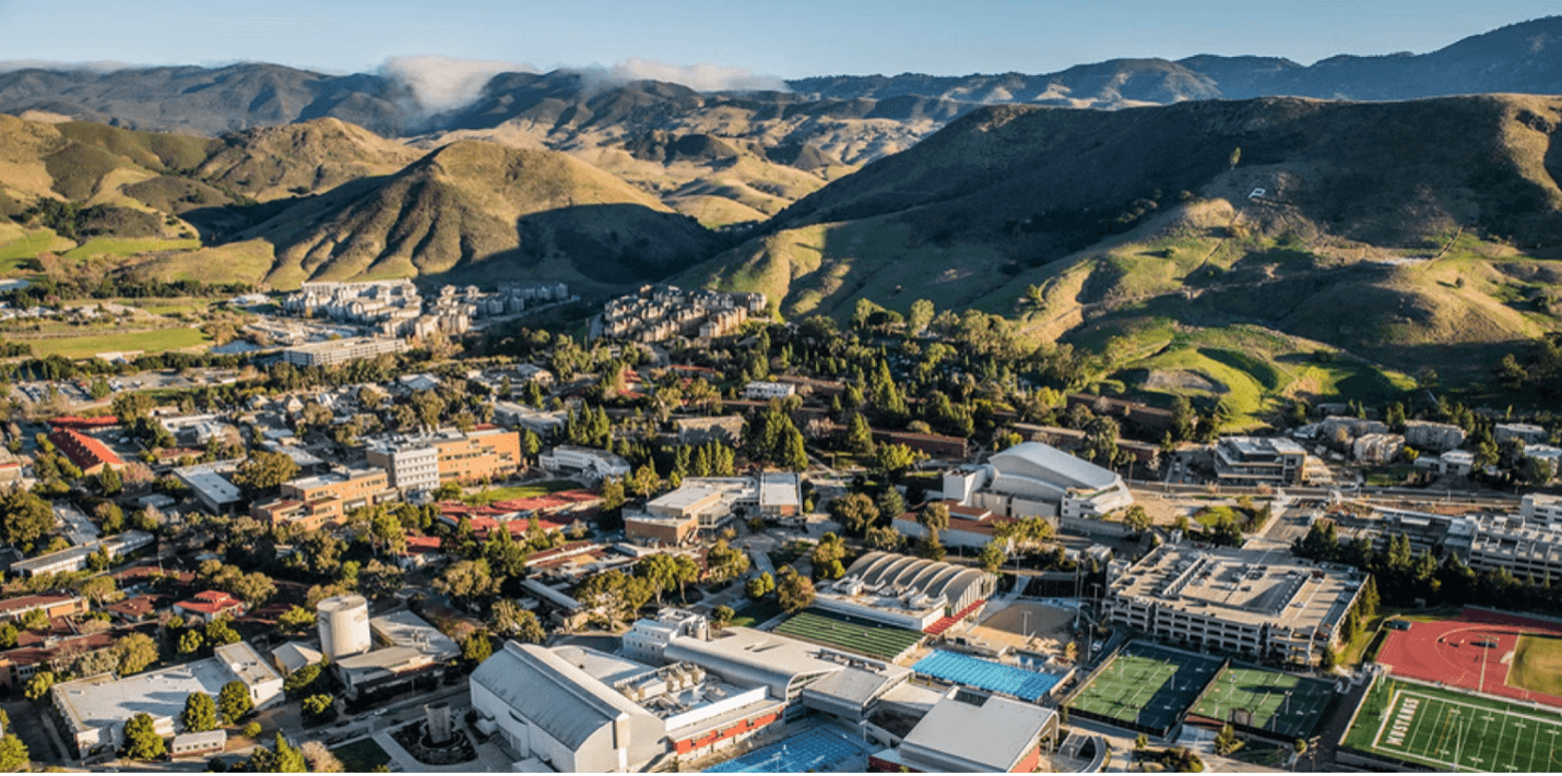 University campus with buildings, sports fields, and tennis courts, surrounded by green hills under a clear sky.