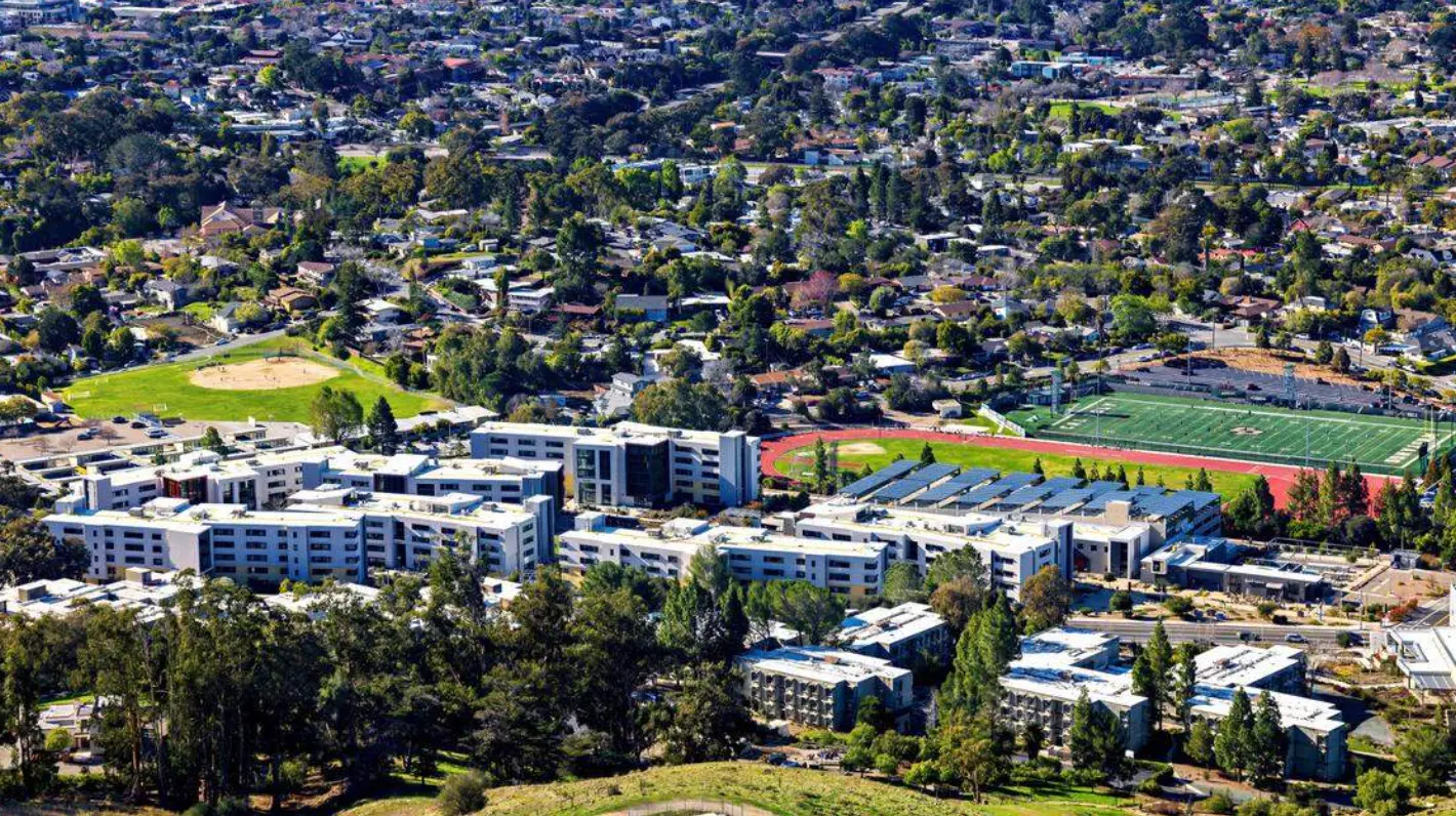 Aerial view of a residential area with apartment buildings, green trees, a football field, and a running track.