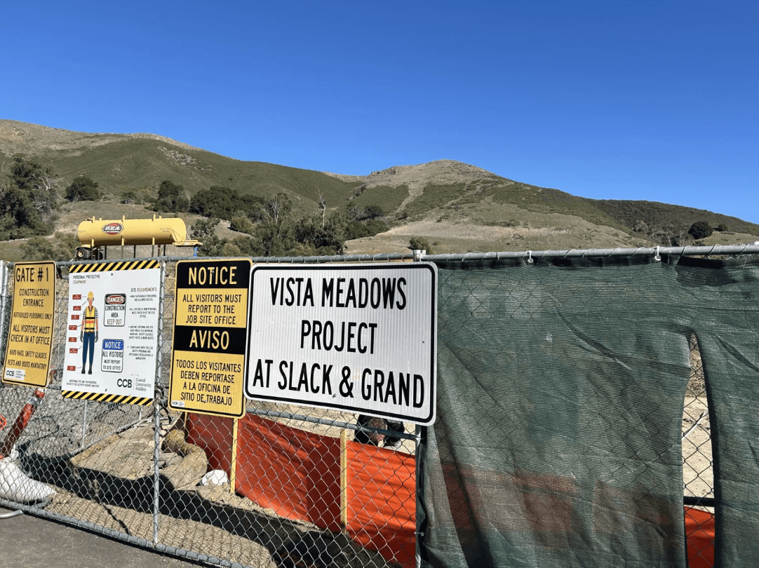 Construction site entrance with multiple warning signs and mountainous landscape in the background.