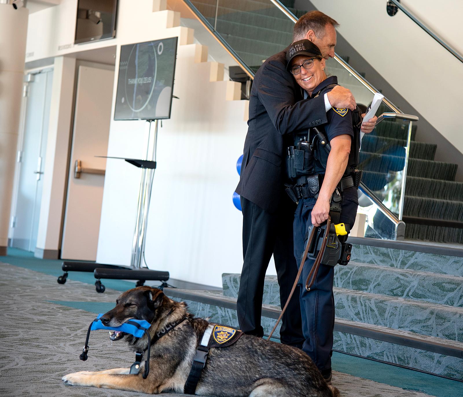 Cal Poly police officer hugs a colleague during a ceremony while a police K-9 wearing a vest lies on the floor chewing a toy.