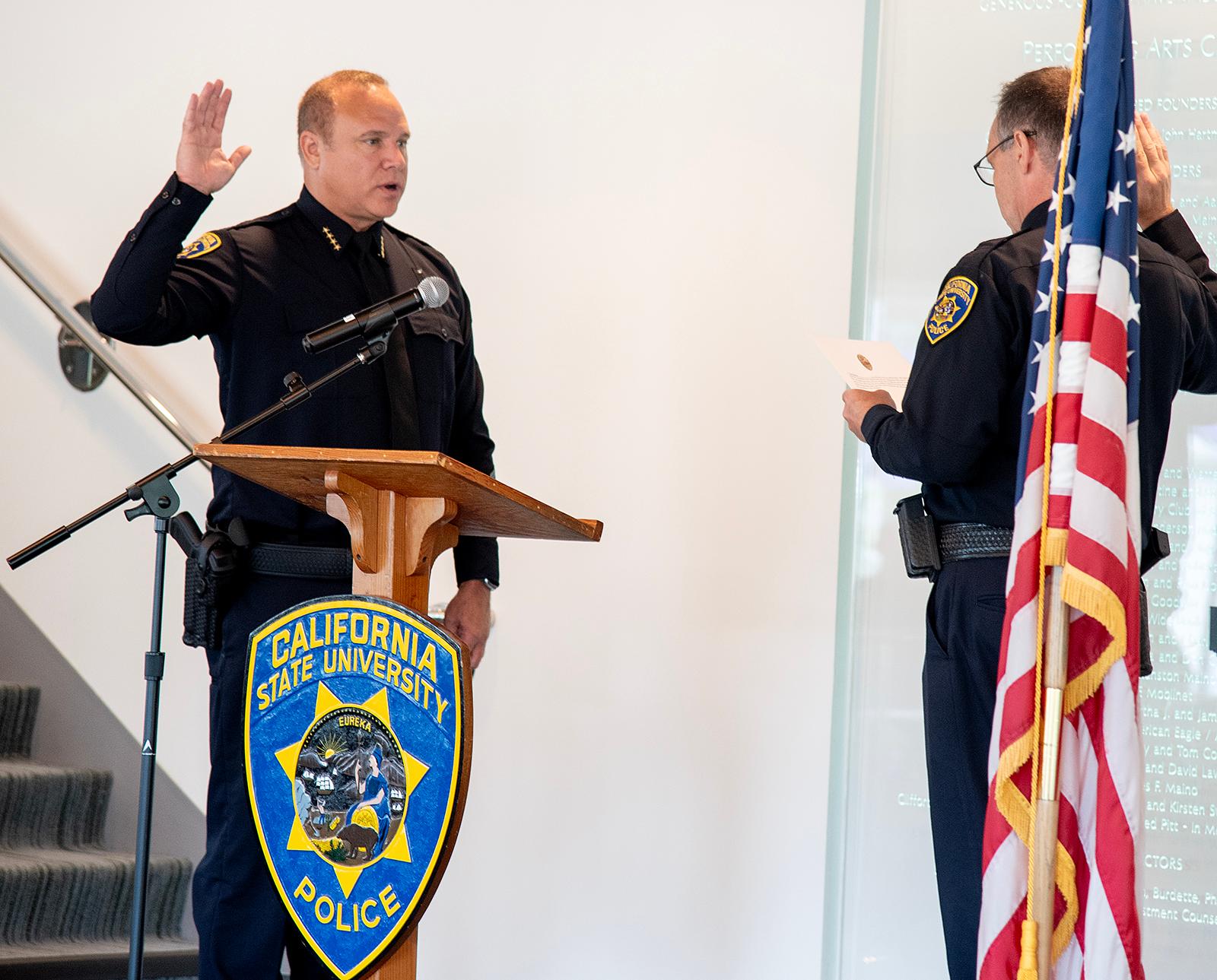California State University police chief stands at a podium with one hand raised, taking the oath of office from another uniformed officer beside an American flag.
