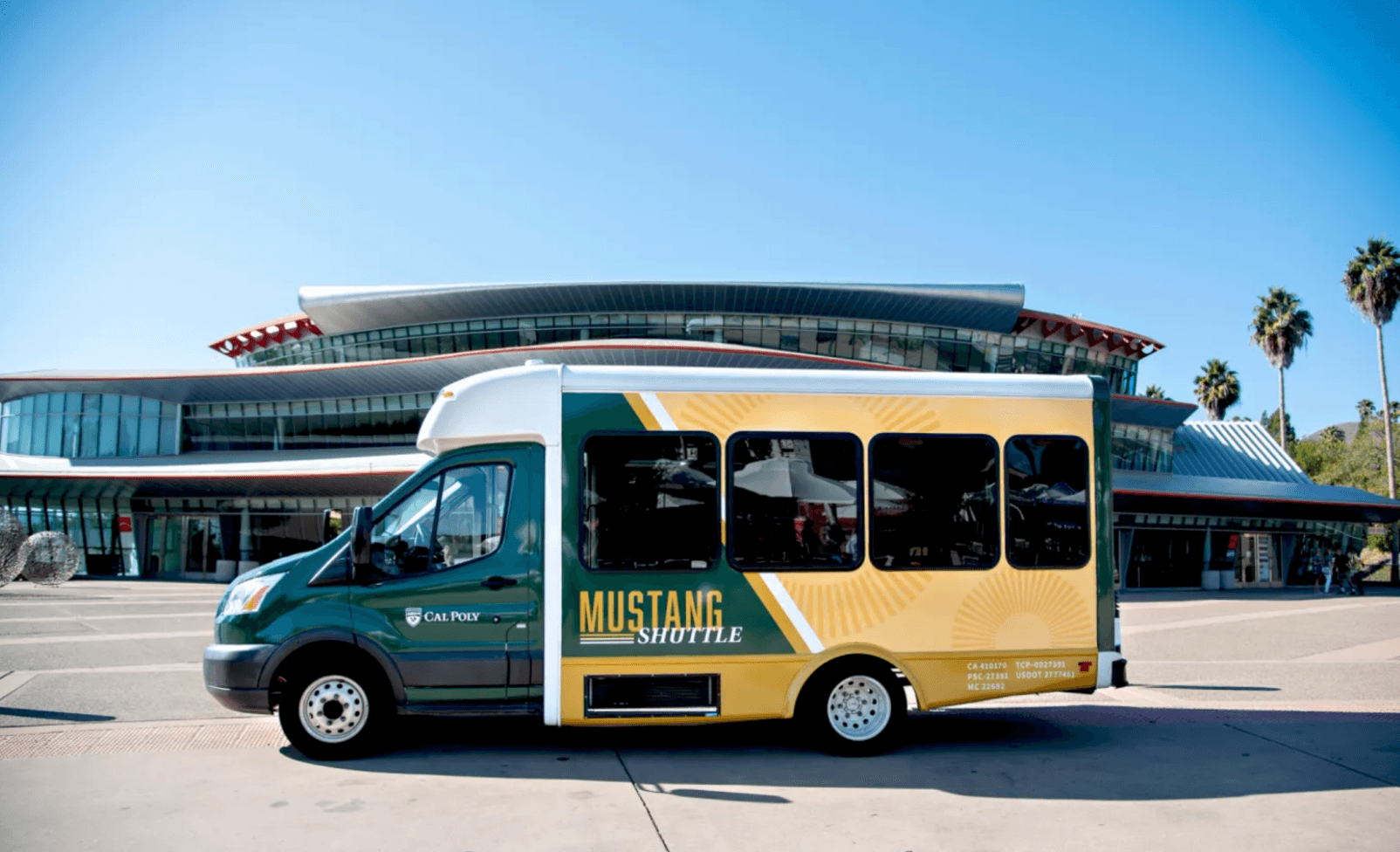 Cal Poly Mustang Shuttle parked in front of the University Union, with the modern glass-and-metal building and palm trees in the background on a clear sunny day.