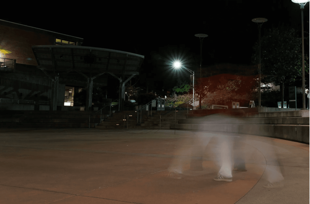 Nighttime photo of an outdoor campus plaza with stairs and overhead lights, showing a faint, ghost-like blur of a person walking due to long-exposure motion.