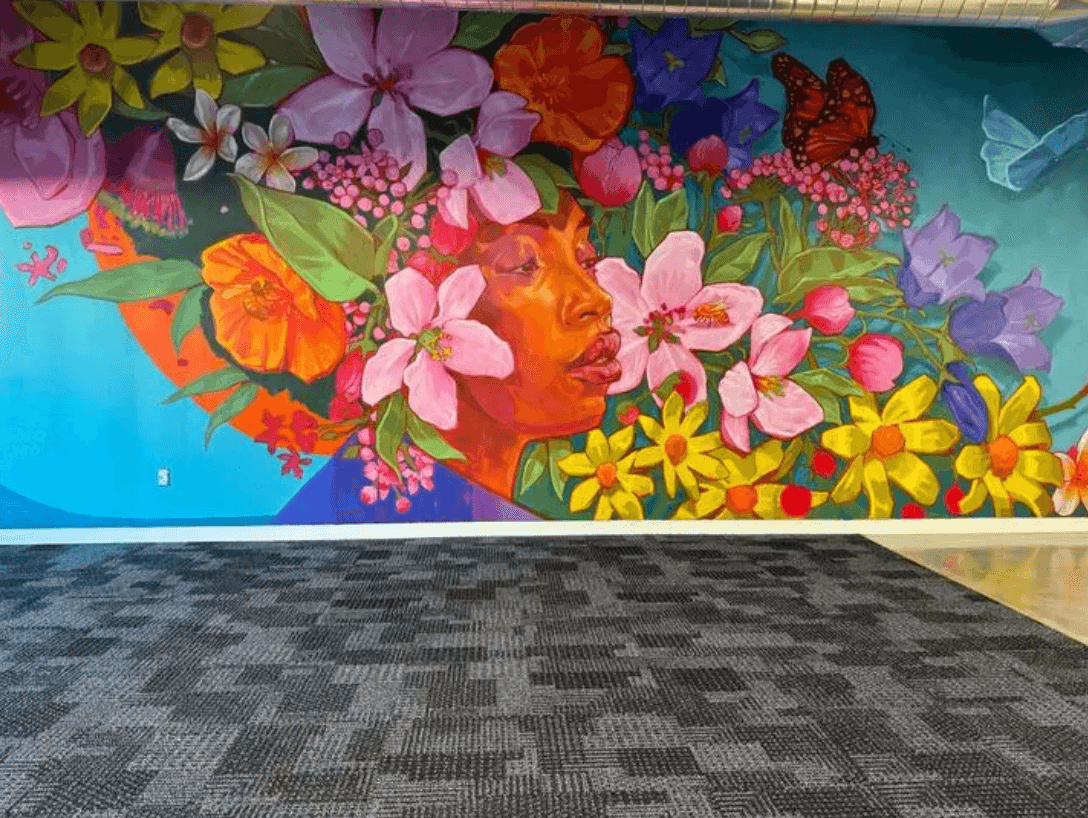 Large indoor mural featuring a woman’s face surrounded by vibrant flowers, leaves, and butterflies painted in bright colors across a blue background.