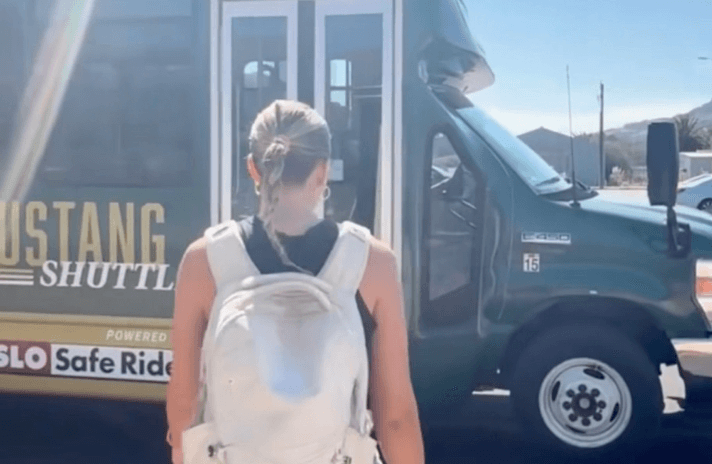 Person with a white backpack walking toward a green Mustang Shuttle bus on a sunny day.